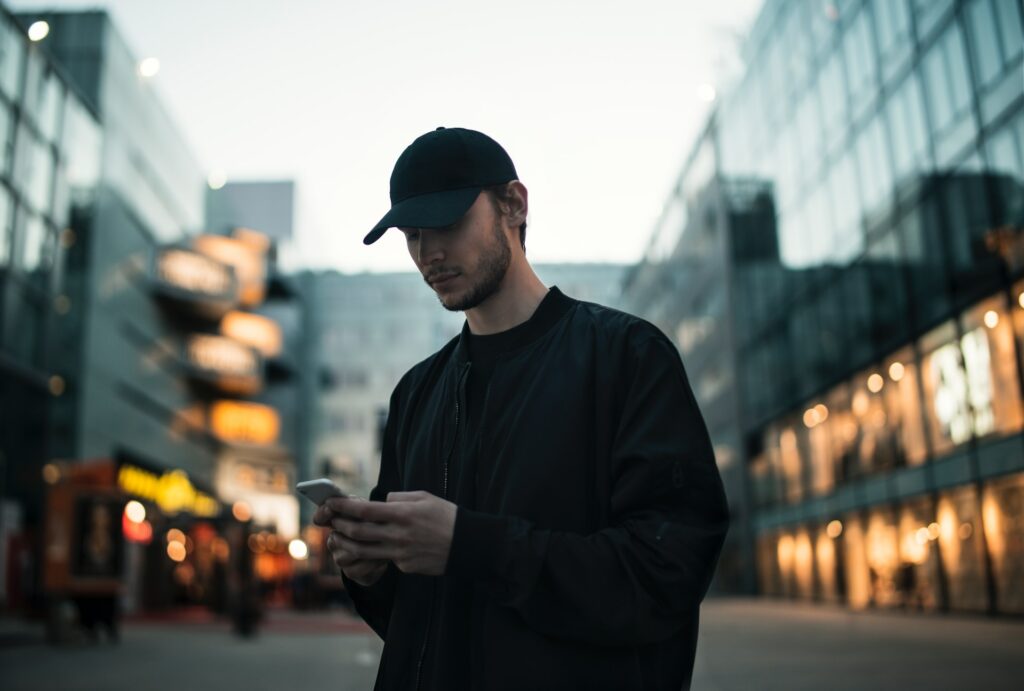 Um homem branco usando uma jaqueta e um boné preto, ele está parado em uma rua mexendo em um celular