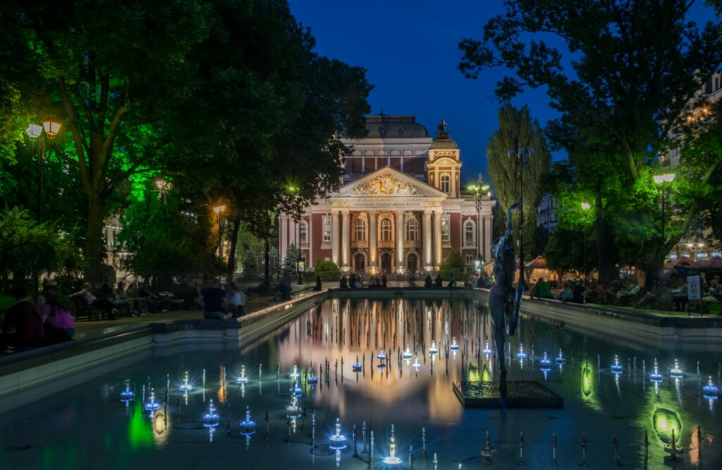 Mansão marrom com uma piscina iluminada para ilustrar o post sobre chip internacional para a Bulgária. A foto foi tirada à noite, e há diversas pessoas sentadas em bancos perto das árvores e ao redor da piscina. - Foto: Valeri Terziyski via Unsplash