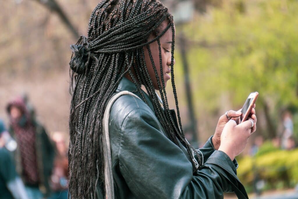 mulher negra vista de perfil enquanto usa o celular, segurando-o com as duas mãos. Ela está em algum parque urbano, pois é possível ver várias árvores e pessoas por perto