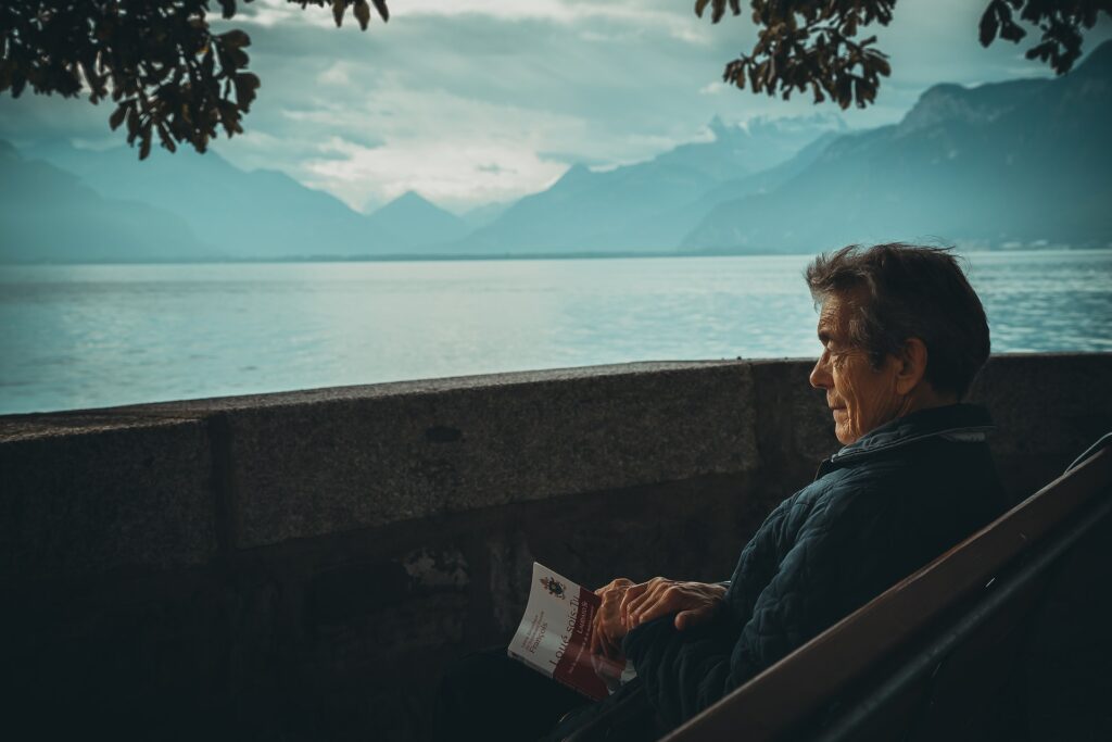 Um homem mais velho sentada de frente para o mar enquanto segura um livro, para representar o seguro viagem para terceira idade
