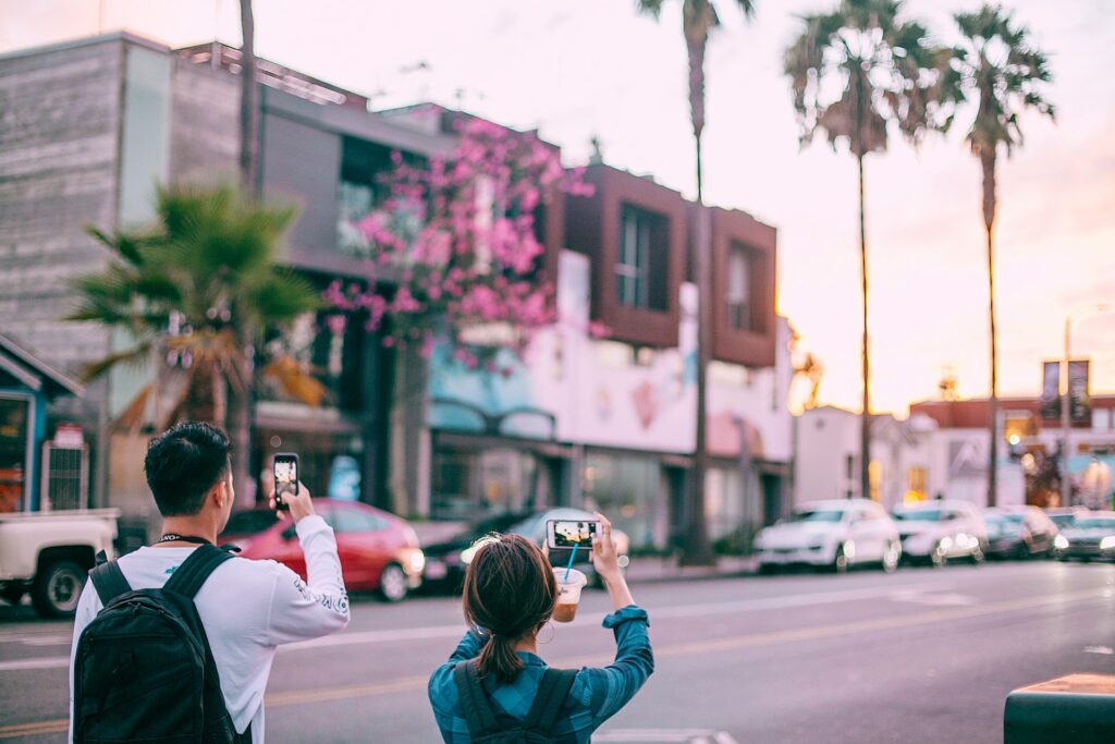 Casal, com mochila nas costas, de costas, tirando foto de uma rua que tem carros estacionados na frente da calçada e um raio de sol alaranjado