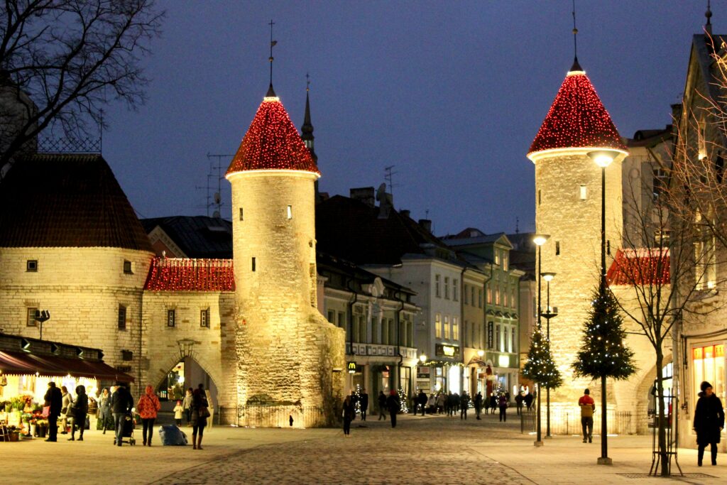 Vista da grandes torres de vigia em mármore construídas no século 14 na entrada da Cidade Antiga de Tallinn durante a noite iluminada com pessoas caminhando no local.
