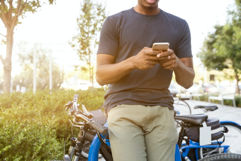 Homem negro, vestido de camiseta cinza e calça bege, encostado em uma bicicleta azul, mexendo no celular com as duas mãos. Ao fundo há natureza e reflexo do sol no lado esquerdo