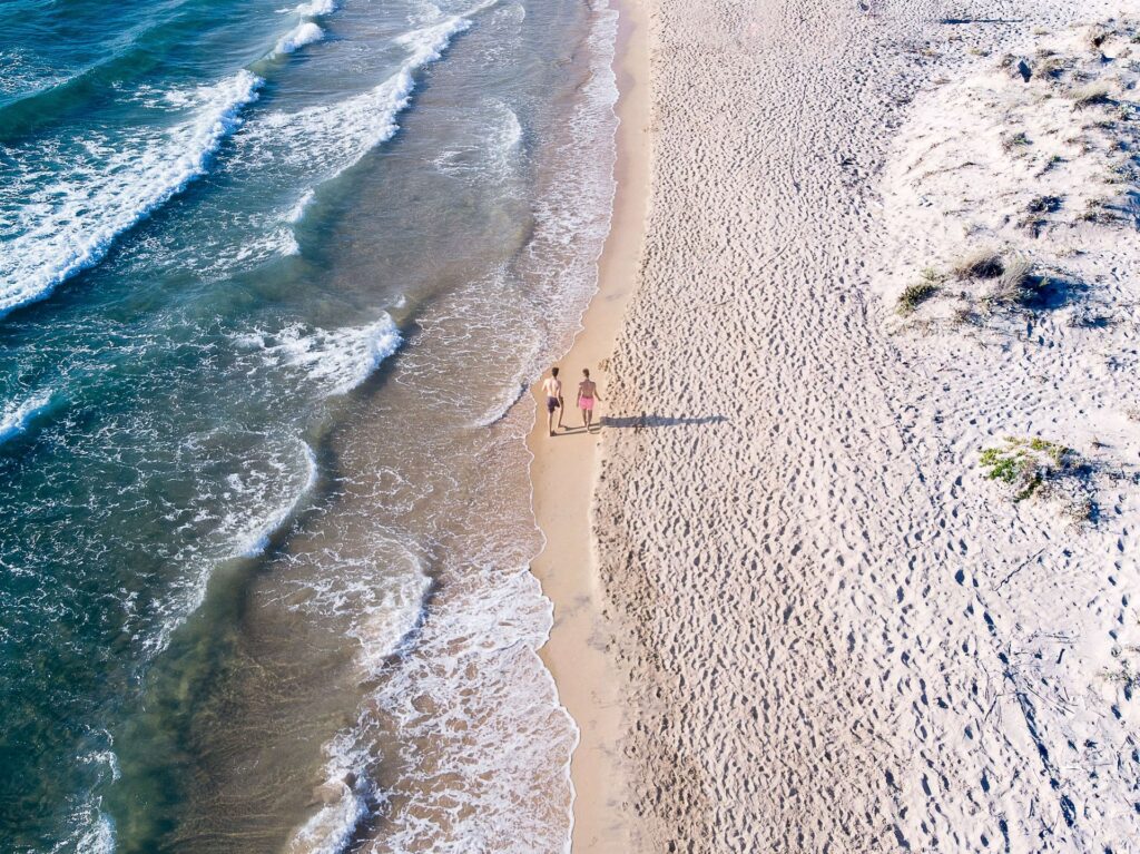 casal visto ao longe em meio a uma praia da Grécia, com águas cristalinas e areia clara, para ilustrar o post de seguro viagem para lua de mel