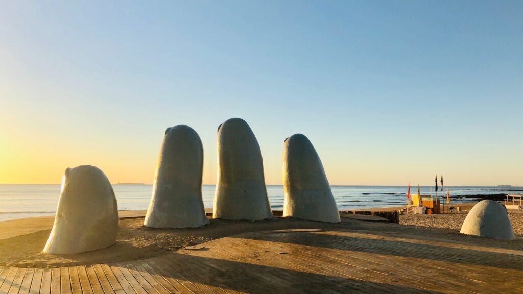 Estátua de dedos saindo do chão para ilustrar o post sobre chip internacional para o Uruguai. Atrás há uma praia, e o sol está se pondo. - Foto: Jared Schwitzke via Unsplash