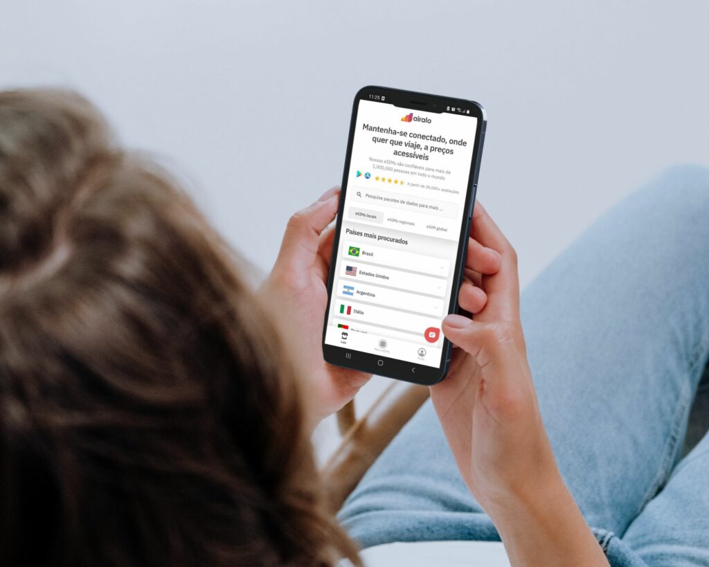Mulher de cabelo castanho mexendo no celular com as duas mãos, vestida de calça jeans, e com a tela do celular no site da Airalo
