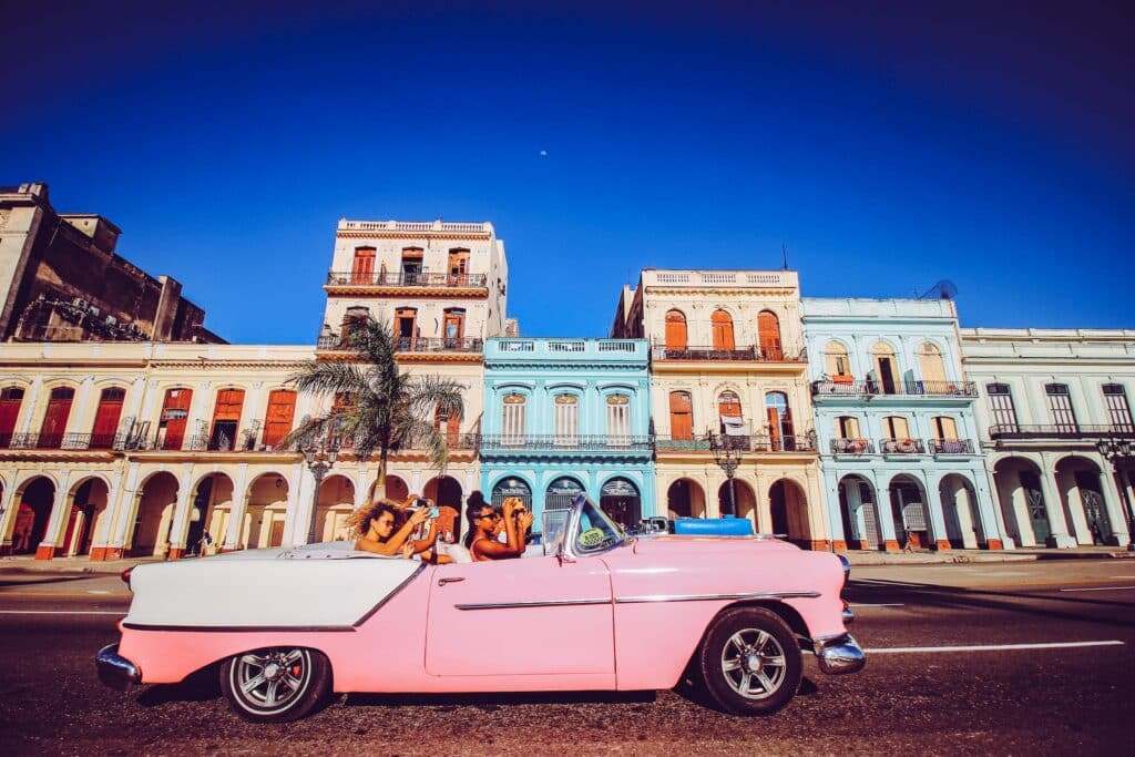 Mulheres tirando fotos com o celular dentro de um carro rosa conversível. a foto foi tirada durante o dia, e o carro está passando na frente de casas beges e azuis, em Cuba. Representa eSIM Cuba.