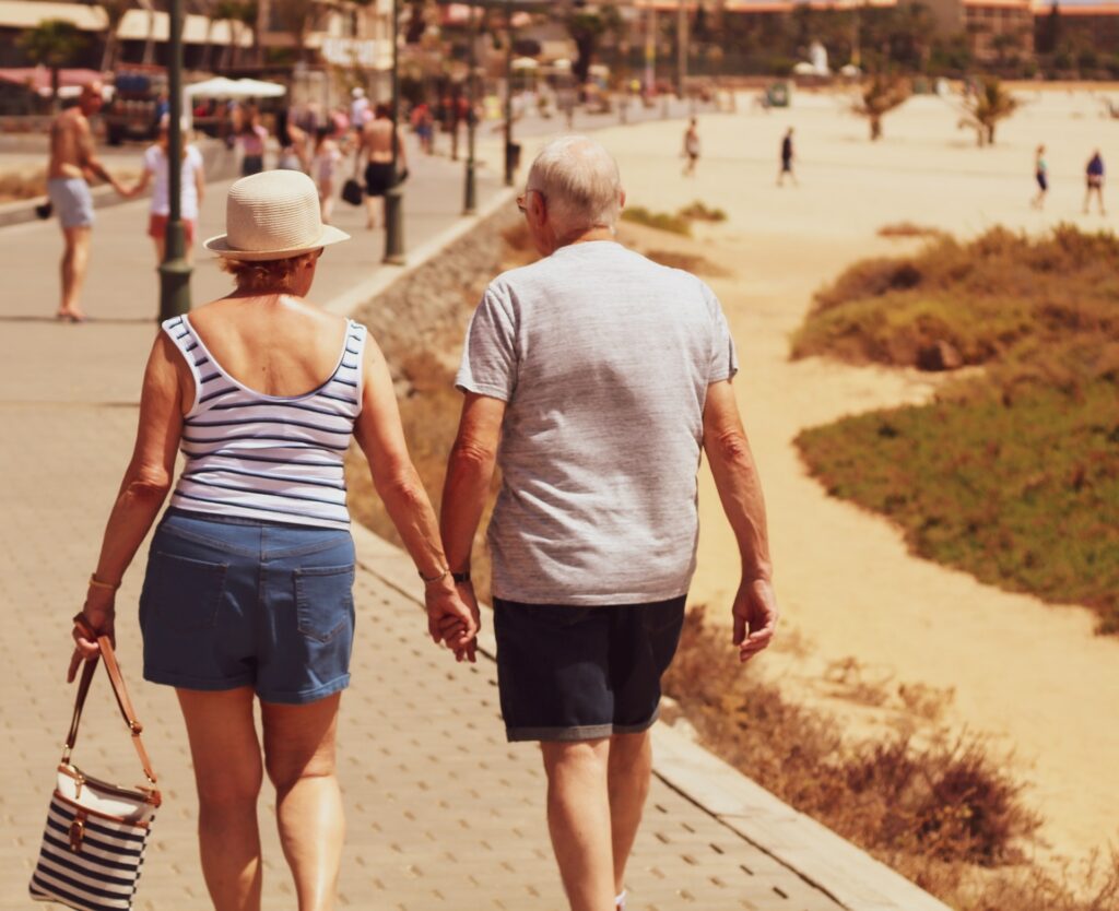 Um casal de idosos de costas, caminhando de mãos dadas na orla asfaltada de uma praia