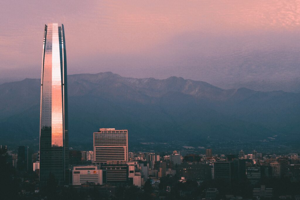 Prédio alto se destacando em meio a outras construções para ilustrar o post sobre chip internacional para Santiago. Há montanhas ao fundo, e a cidade está encoberta por uma neblina durante o por do sol rosa e roxo. - Foto: Agustin Ljosmyndun via Unsplash
