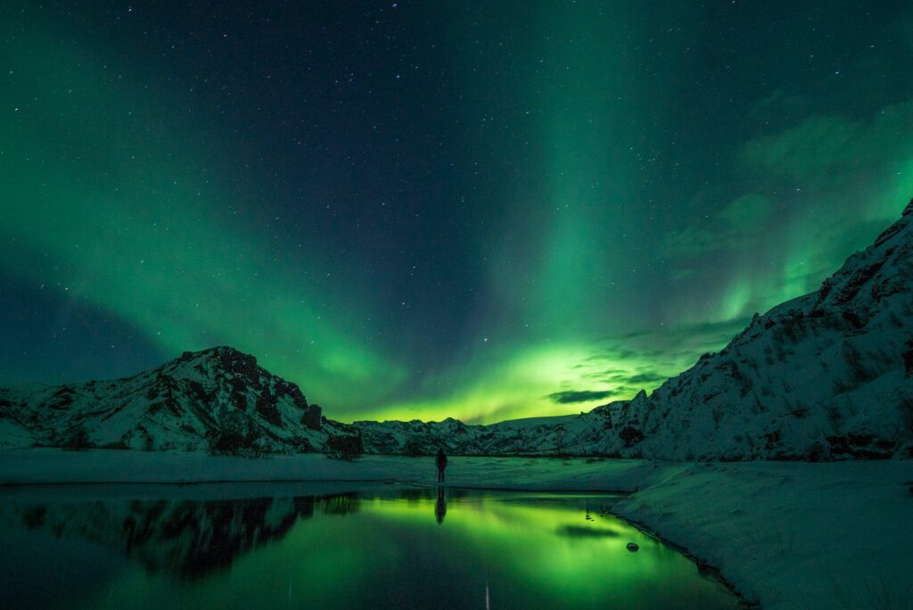 Pessoa no meio de grandes montanhas cobertas de neve e um lago refletindo a cor verde da aurora boreal, ilustrando post chip internacional para a Islândia.