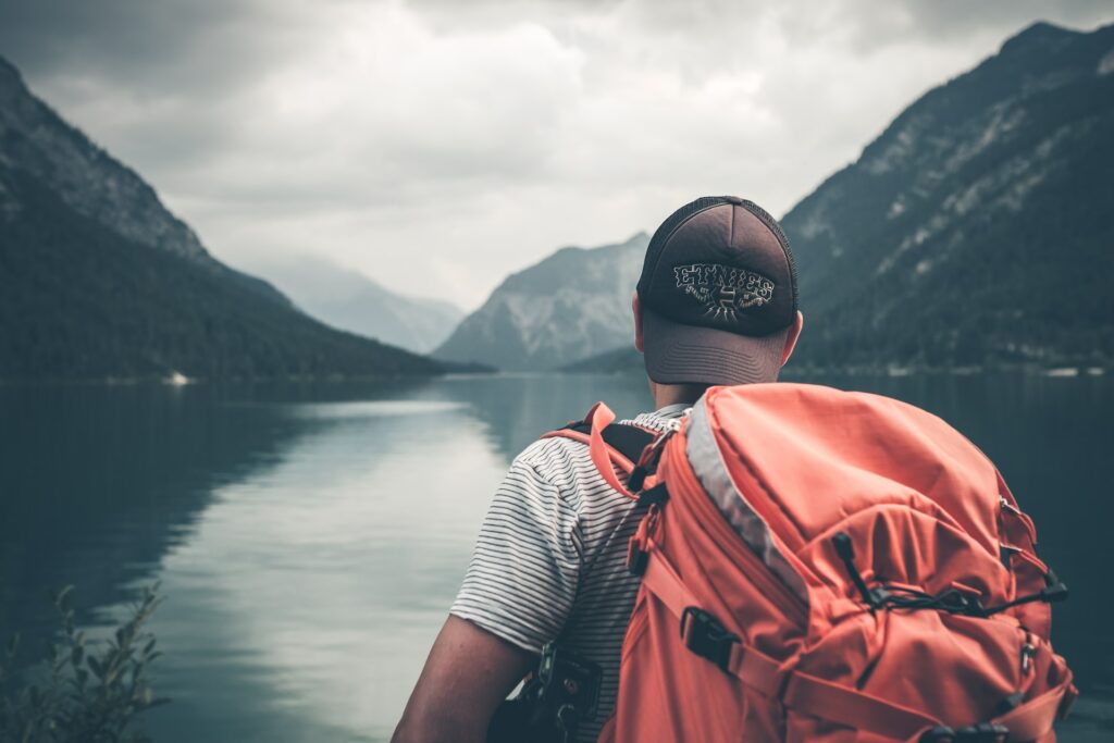 homem com mochila laranja grande nas costas na frente de um lago com águas plácidas e montanhas em volta, na Áustria