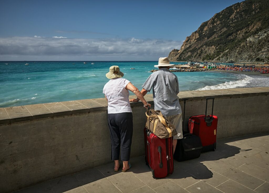 Dois idosos segurando malas de viagem sob uma pequena passarela que dá vista para o mar, os dois estão observando a praia