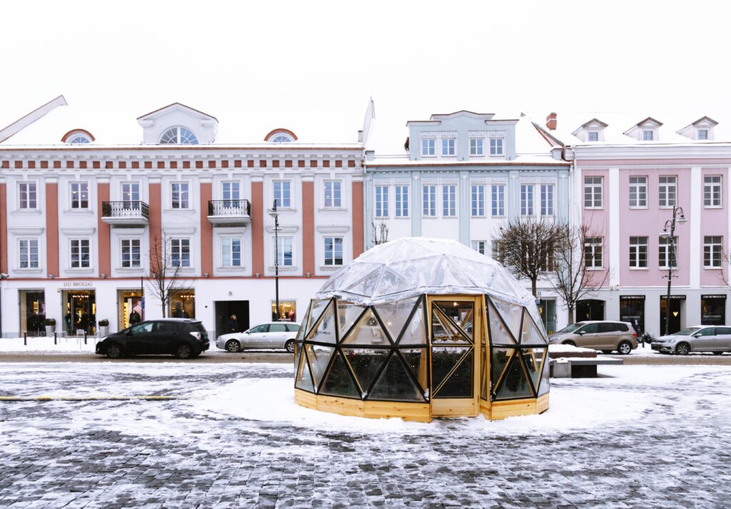 Vista da cidade de Vilnius, Lituânia durante o dia com uma cúpula grande de acrílico no centro, ao fundo os carros e prédios e neve em volta.