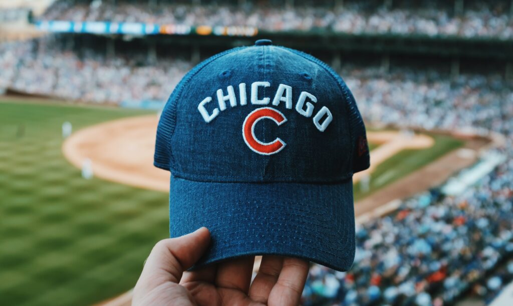 Mão branca segurando um boné de beisebol escrito "Chicago C", em um campo de beisebol. No fundo, vemos uma parte do campo e as arquibancadas desfocadas. Foto para ilustrar o post sobre chip internacional para Chicago.