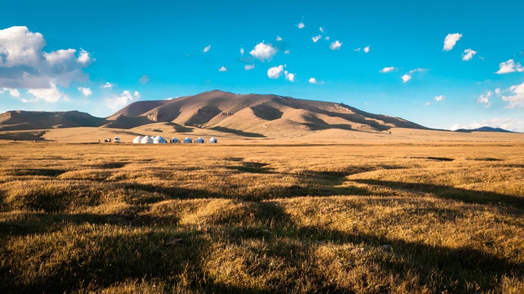 O céu está azul com algumas nuvens, vemos algumas cordilheiras no fundo e yurtas (acomodações nômades). O solo é uma baixa relva. Foto para ilustrar o post sobre chip internacional para o Quirguistão.