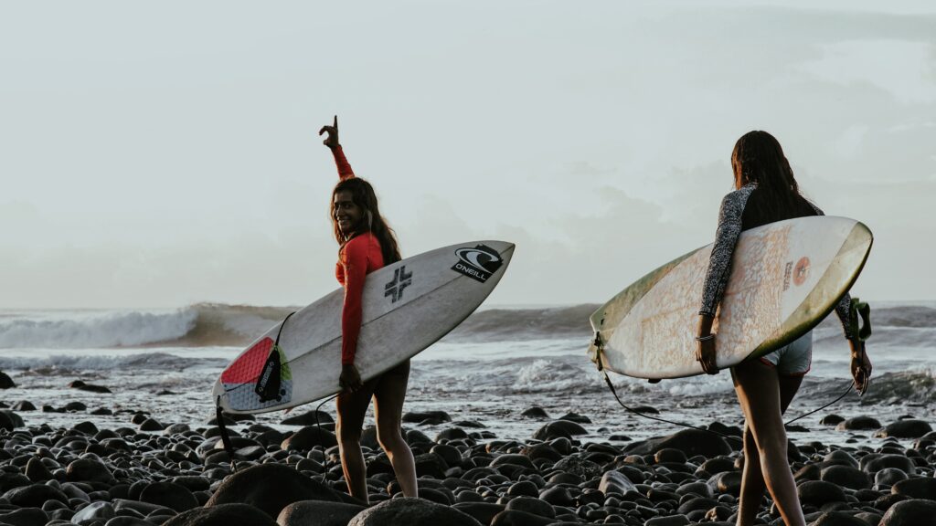 Duas mulheres surfistas segurando uma prancha de surf, caminhando pelas pedras em direção ao mar. O céu etá nublado. Uma delas está acenando para a câmera. Foto utilizada para ilustrar o chip internacional para El Salvador.