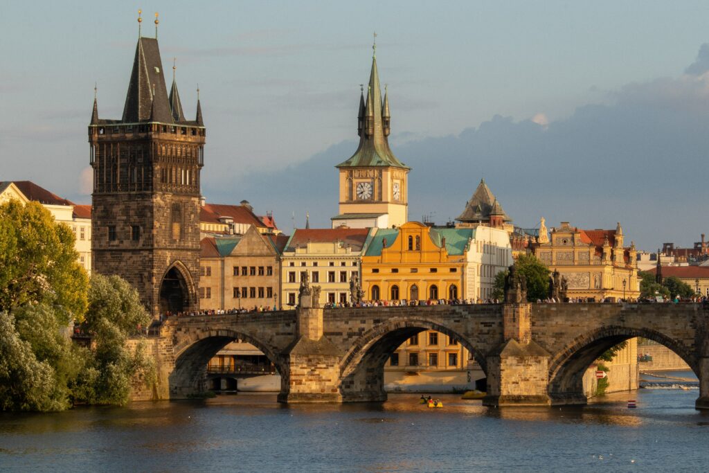 Famosa ponte Charles situada em Praga, na Tchéquia. Vemos o rio abaixo dela, à esquerda, algumas árvores. Atrás, temos a visão de várias construções no estilo europeu e um torre de relógio. Foto para ilustrar o post sobre chip internacional para o Leste Europeu.