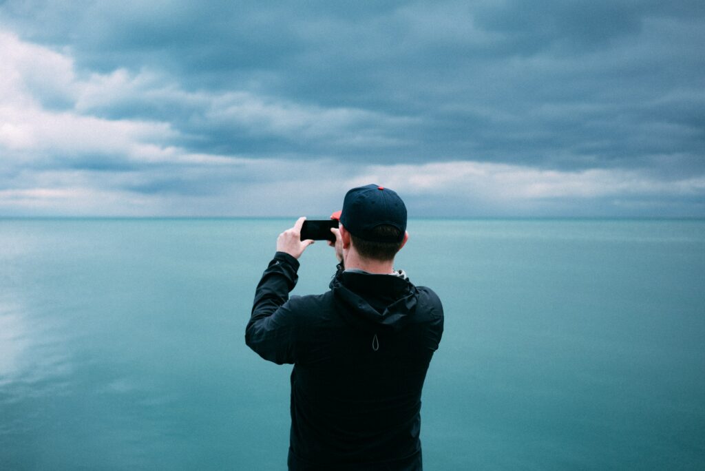 Homem branco, com vestes pretas e um boné preto, tirando foto do mar em uma praia de Chicago. Foto para ilustrar o post sobre chip internacional para Chicago.