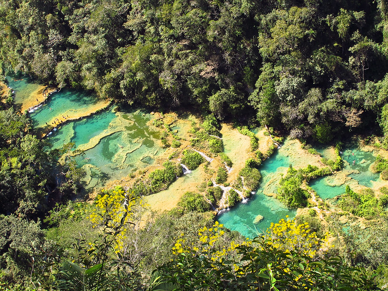 A imagem demonstra a vista aérea do Semuc Champey, beleza natural de Guatemala que mostra as piscinas naturais do país. Há alguns banhistas nelas. Ao redor das águas, há uma vasta vegetação. Imagem para ilustrar o post de chip internacional para Guatemala.