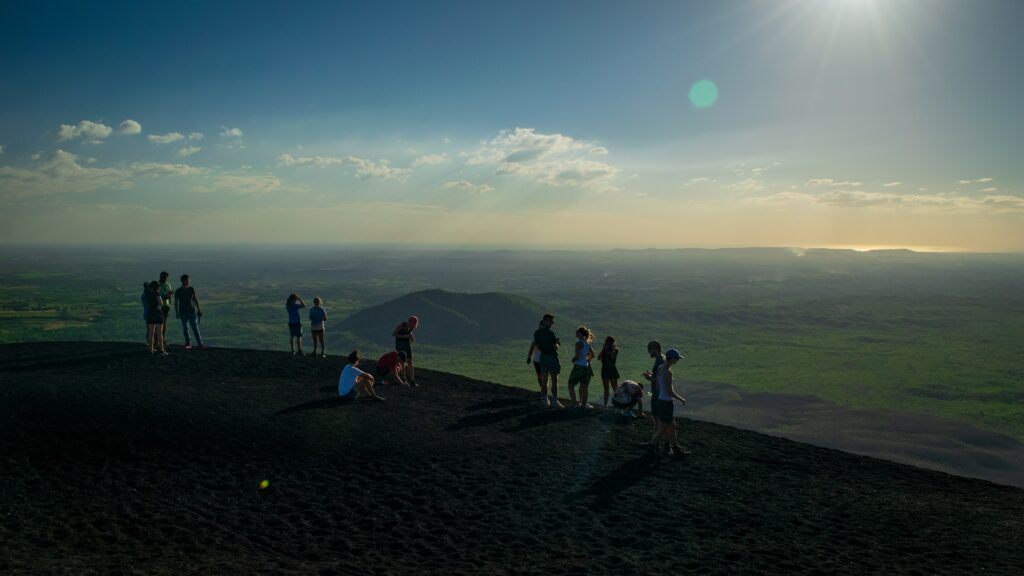 Vista de cima de vários turistas fazendo trilha no Vulcão Cerro Negro, Nicarágua. Está de dia e os viajantes observam o horizonte e conversam entre si. No segundo plano da imagem há uma vasta vegetação. Foto para ilustrar o chip internacional para a América Central.