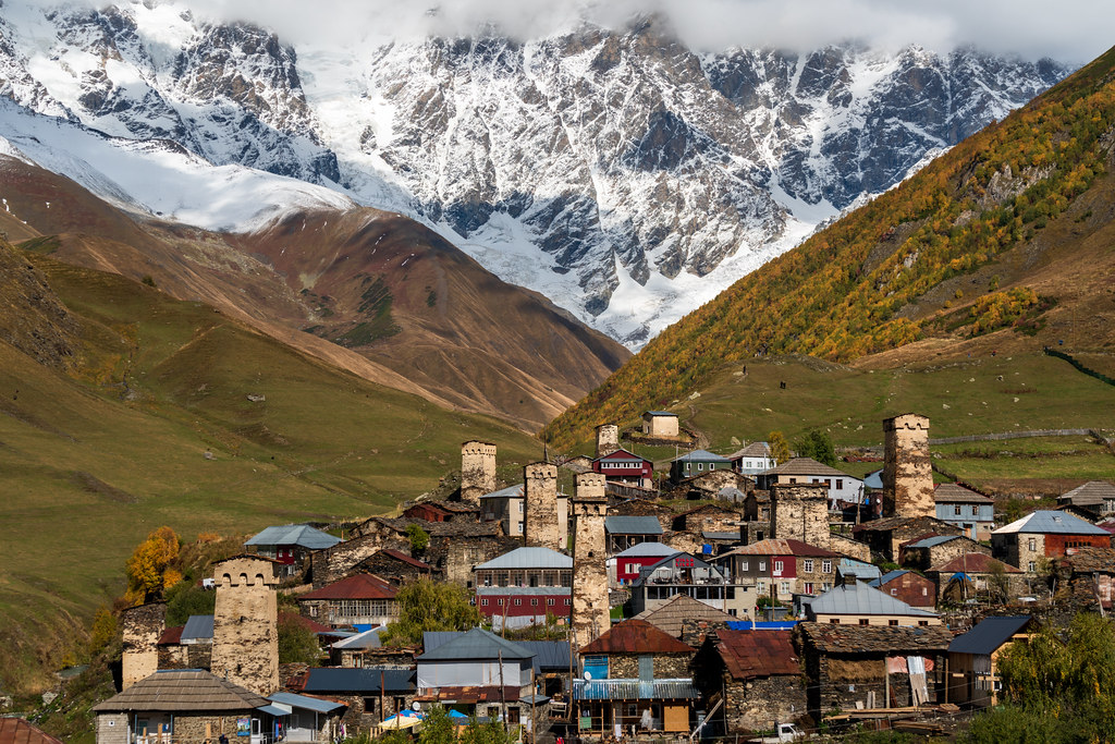 Diversas casas pequenas e coloridas no meio. Ao redor montanhas e atrás montanhas com neve.