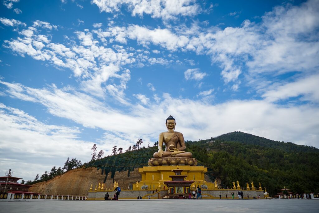 Estátua de Buda ao centro com pessoas em volta e vasta vegetação em segundo plano sob o céu azul com algumas nuvens para representar o chip internacional para o Butão. 