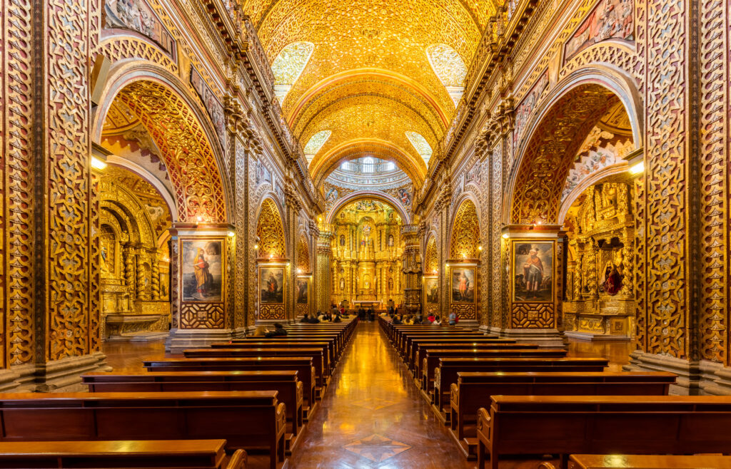 Interior de uma igreja com decoração predominantemente dourada, bancos de madeira e colunas com quadros religiosos.