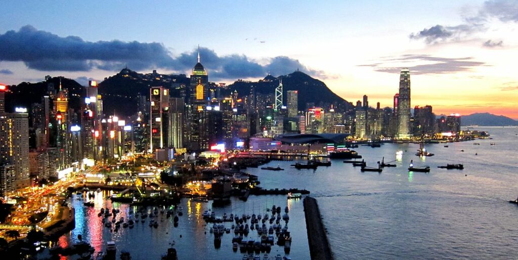 Vista do entardecer, de Hong Kong. Ao lado direito o mar, com vários barcos parados. Ao lado esquerdo uma rua paralela iluminada, com vários carros, a cidade de Hong Kong com prédios iluminados e várias cores.