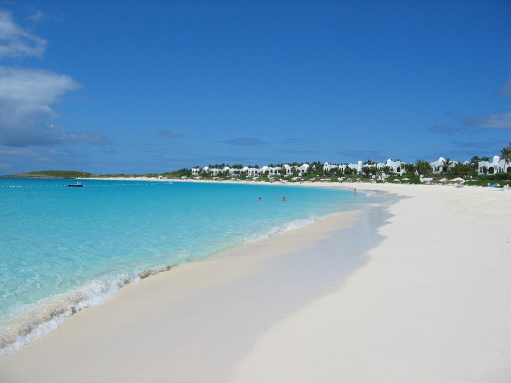 Uma ilha de dia, ao lado direito a areia e ao lado esquerdo o mar, azul claro. No fundo algumas árvores e casas