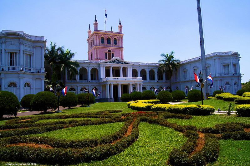 Palácio branco e rosa, com várias árvores ao redor. Em frente há um grande jardim e pequenas bandeiras do Paraguai. Está de dia. Foto para ilustrar post chip internacional para Assunção.