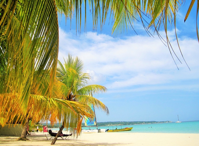 Pessoa deitada em uma cadeira na praia e olhando o mar em frente. Algumas palmeiras e outras pessoas estão na areia também e há um barco na água. Imagem para ilustrar post sobre chip internacional para a Jamaica.