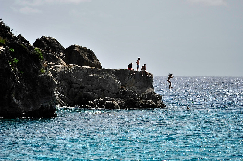 Pessoas pulando de uma pedra alta na beira do mar azul durante o dia.