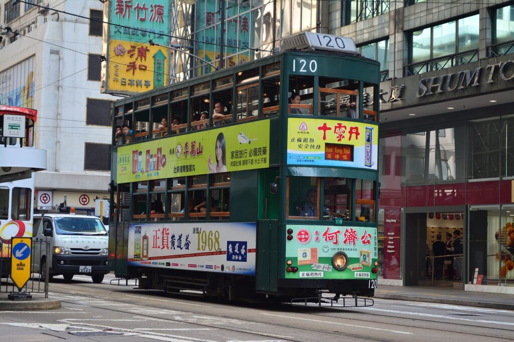 Trem verde de Hong Kong passando no meio da rua, ao redor  prédios com lojas. Atrás do trem, um carro branco.