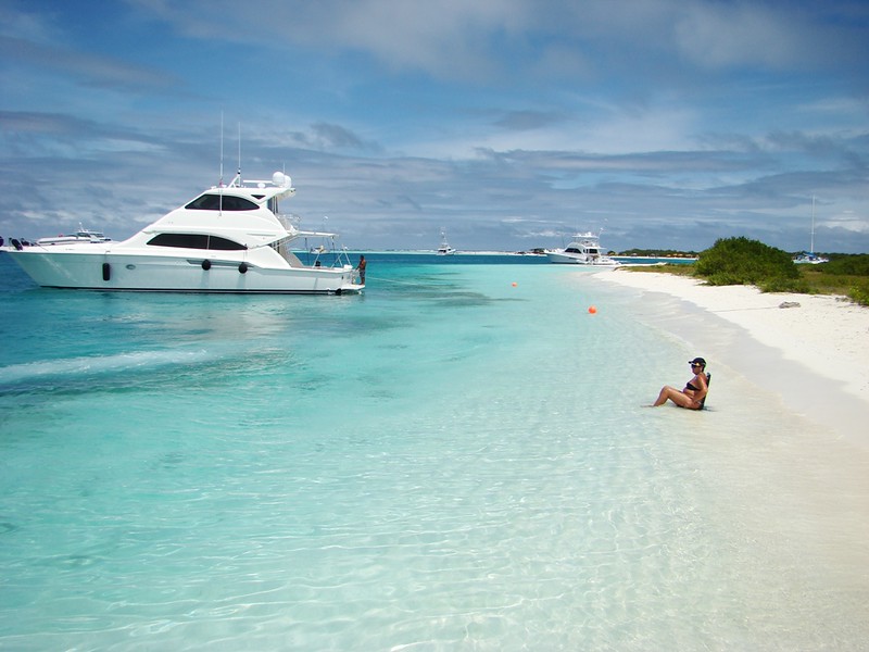 Foto de praia com uma mulher sentada na beira do mar e barcos ao fundo. Está de dia, com um céu bem azul. Foto para ilustrar post chip internacional para Los Roques.