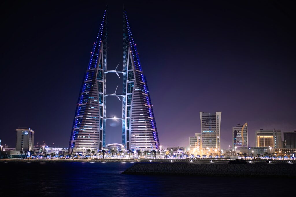 Duas torres com formato triangular ligadas por 3 pontes de aço, e ao lado da torre outras construções iluminadas durante a noite, ilustrando post chip internacional para o Bahrein.