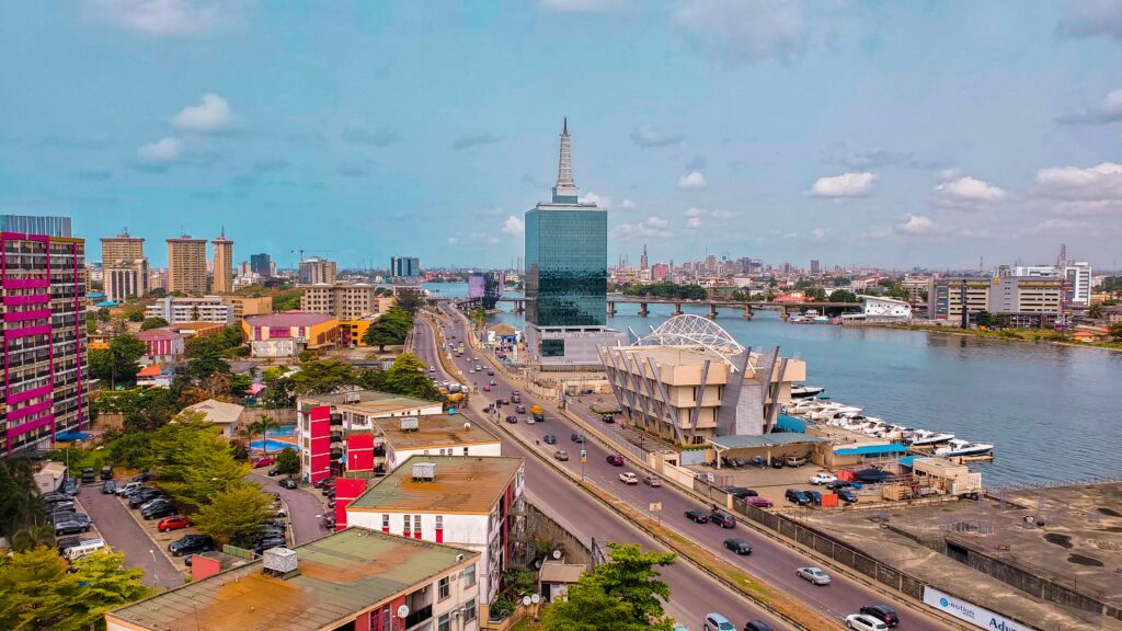 Foto da cidade de Lagos, Nigéria durante o dia com casas, prédios no lado esquerdo da imagem, no centro tem uma rodovia e do lado direito um rio. Representa seguro viagem para a Nigéria.