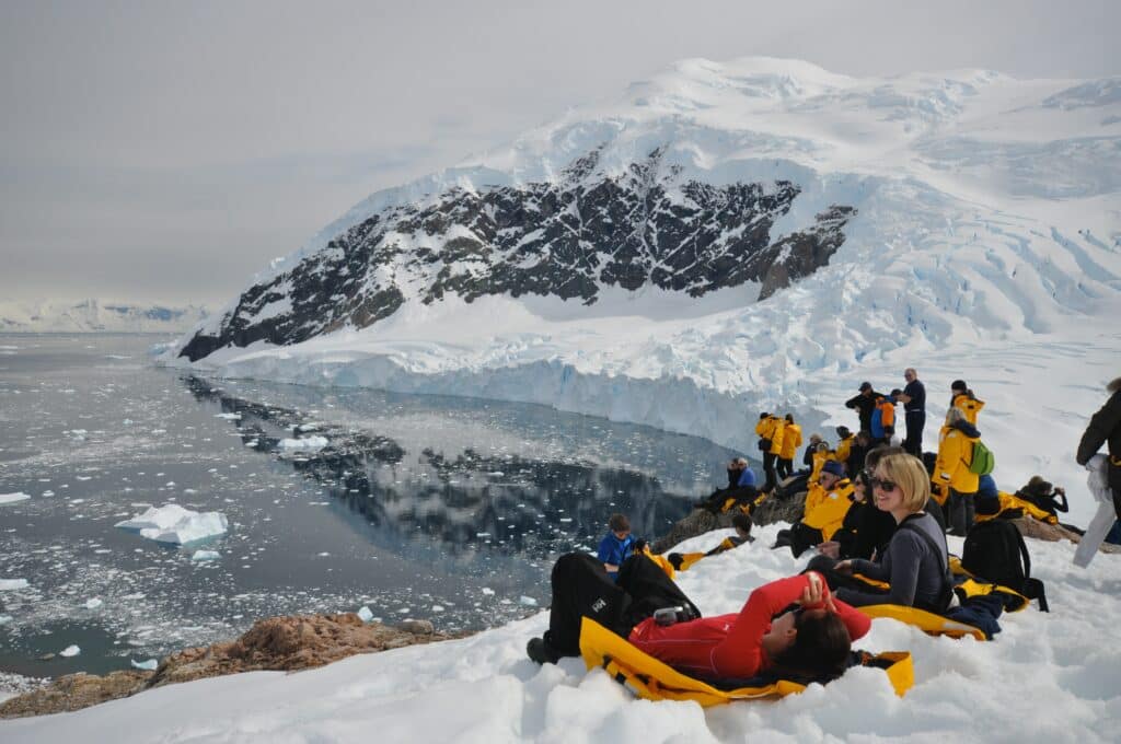 Um grupo de pessoas do lado direito da imagem sentados na neve na Antártida em frente ao um lago.