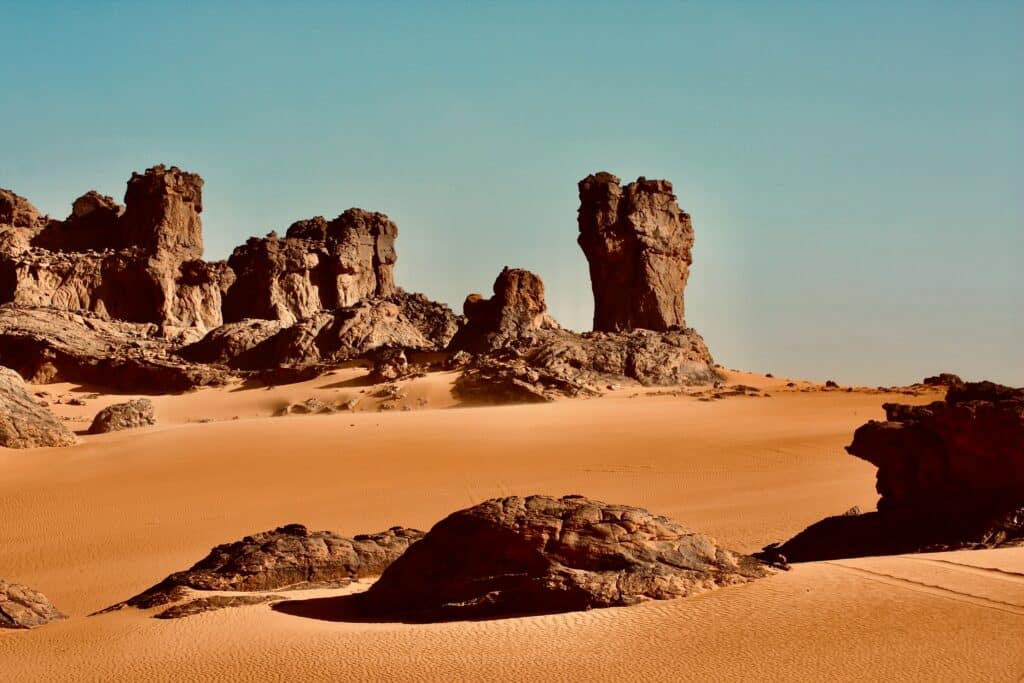 Imagem do Tassili du Hoggar, In Guezzam na Argélia com deserto, a frente e ao fundo rochas durante o dia. Representa seguro viagem para a Argélia.