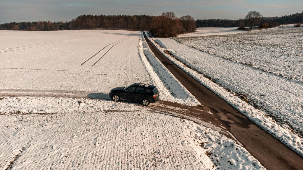 Vista aérea de um carro preto dirigindo em uma estrada estreita coberta de neve em Mühlhausen através de extensos campos nevados com uma fileira de árvores ao fundo. Representa aluguel de carros na Alemanha.