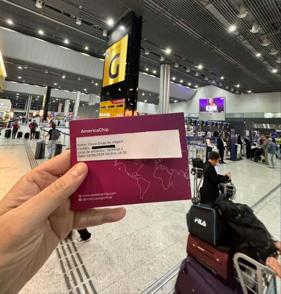Uma pessoa segura um cartão eSIM da America Chip no terminal do aeroporto de guarulhos com viajantes e bagagens ao fundo.