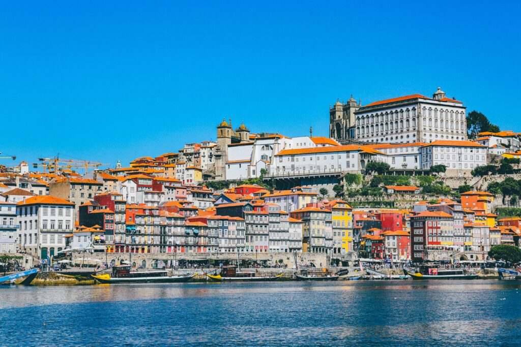 Uma paisagem urbana à beira-mar no Porto com edifícios coloridos e uma estrutura proeminente no topo de uma colina sob um céu azul claro. Representa chip internacional para o Porto.