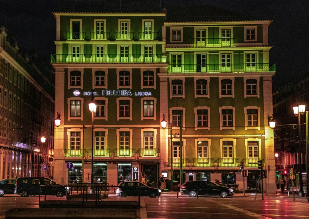 Vista noturna de um hotel de vários andares iluminado com iluminação verde, carros na frente e uma rua com postes de luz e pedestres em Lisboa. Representa aluguel de carros em Lisboa.