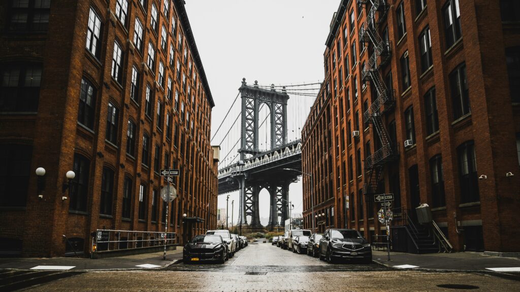 Uma rua de Dumbo entre prédios de tijolos leva a uma vista da Ponte de Manhattan em um dia nublado. Carros estão estacionados ao longo da estrada. Representa aluguel de carros em Nova York.