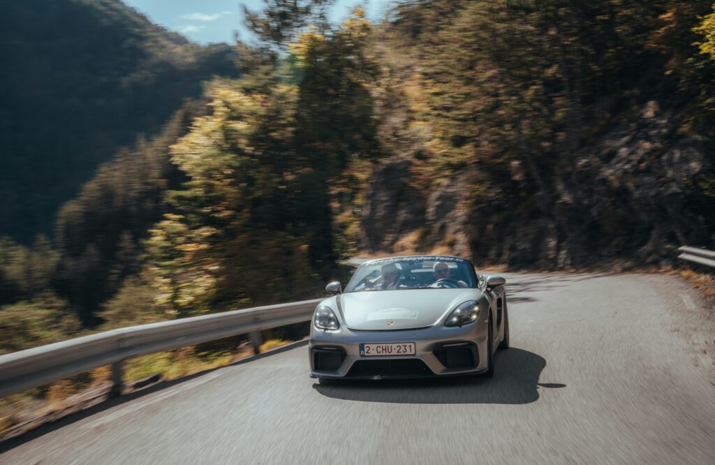 Um carro esportivo prateado dirige em uma estrada sinuosa na montanha dos Alpes, cercado por árvores, sob um céu azul claro. Representa aluguel de carros na França.
