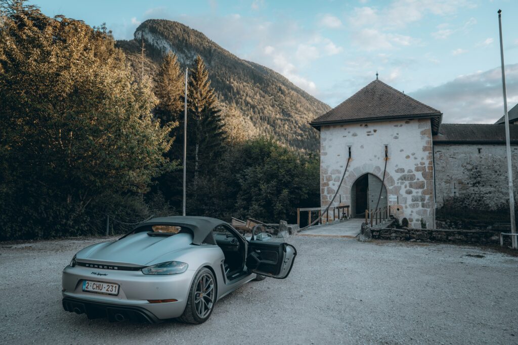 Um carro conversível prateado com a porta aberta está estacionado em frente a um edifício de pedra com uma entrada em arco, perto de uma montanha nos Alpes. Representa aluguel de carros na França.