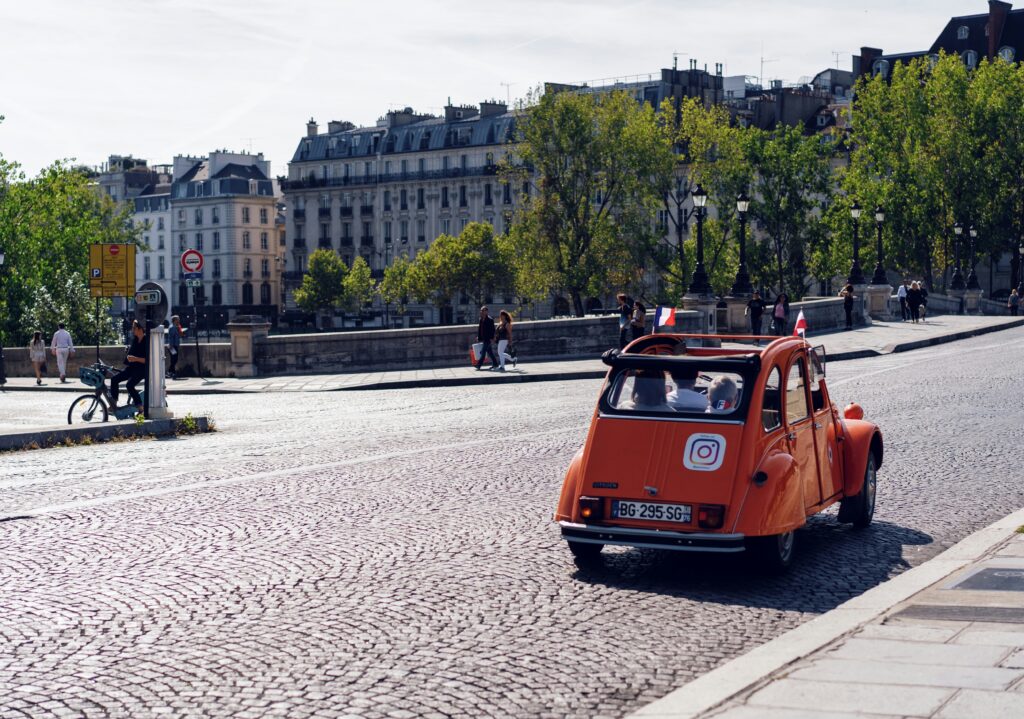 Um carro antigo laranja circula por uma rua de paralelepípedos em Paris, com pessoas caminhando e prédios ao fundo. Representa aluguel de carros em Paris.