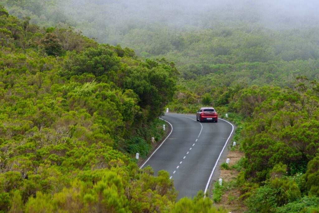 Um carro vermelho dirige em uma estrada sinuosa em Fanal, através de colinas verdejantes sob um céu enevoado. Representa aluguel de carros em Portugal.