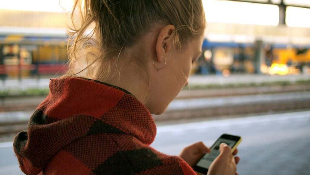 Mulher de cabelos loiros verifica seu telefone em pé na plataforma de um trem, vestindo uma jaqueta xadrez vermelha e preta.