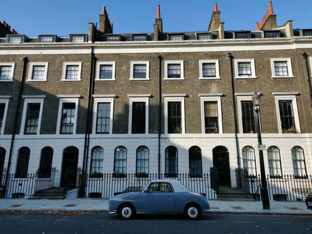 Uma fileira de sobrados em estilo georgiano com um carro azul antigo estacionado na rua em frente a eles, sob um céu azul claro. Representa aluguel de carros em Londres.