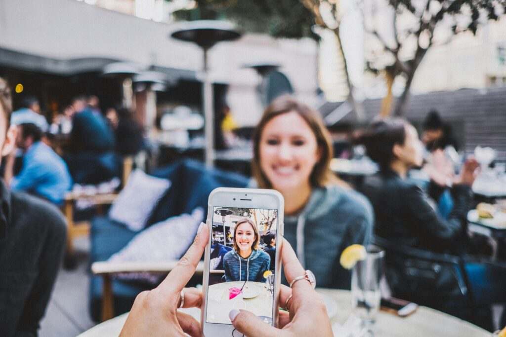 Uma pessoa segura um telefone e captura uma mulher sorridente sentada em um café ao ar livre, com pessoas e mesas ao fundo.